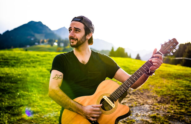 Der Singer/Songwriter Aljosha Konter (Mann mit Gitarre vor grüner Wiese mit Bergen im Hintergrund)