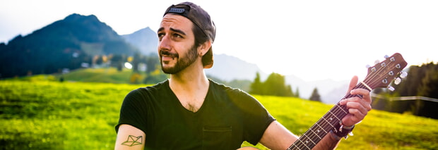 Der Singer/Songwriter Aljosha Konter (Mann mit Gitarre vor grüner Wiese mit Bergen im Hintergrund)