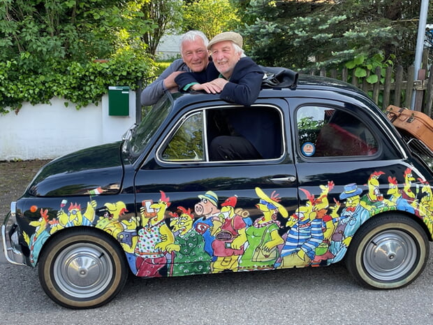 Cartoonist Peter Gaymann und Musik-Kabarettist Josef Brustmann schauen aus einem Auto mit Hühner-Cartoons von Peter Gaymann.