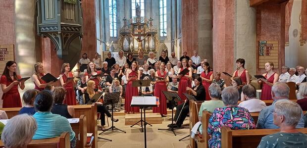 Chor singt in einer Kirche. Menschen sitzen und stehen während der Aufführung zuhörend da. Hier zu sehen: Chor der Regiswindiskirche (sitzend im Hintergrund), Vokalensemble Novere (im Vordergrund, rot gekleidet), Streicherinnen. - Aufnahme aus dem Konzert "Hörst Du nicht die Bäume rauschen?" vom 19.7.2026 unter der Leitung von Katharina Linn.