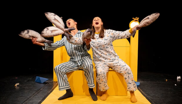 Szenenfoto aus "Hey, hey, hey, Taxi!" gespielt von der Badischen Landesbühne. Zu sehen sind 2 Personen auf einem großen gelben Bett in Schlafanzügen, ein Mann und eine Frau., die fröhlich singen und gestikulieren.