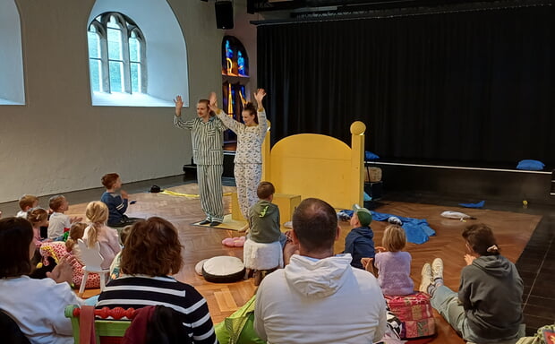Szenenfoto aus der Aufführung der Badischen Landesbühne von "Hey, hey, hey, Taxi!": Zwei Personen in Schlafanzügen stehen vor Kindern auf einer Bühne. Publikum (von hinten sichtbar) sitzt im Kreis.