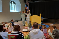 Szenenfoto aus der Auff&uuml;hrung der Badischen Landesb&uuml;hne von "Hey, hey, hey, Taxi!": Zwei Personen in Schlafanz&uuml;gen stehen vor Kindern auf einer B&uuml;hne. Publikum (von hinten sichtbar) sitzt im Kreis. 

