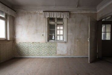Blick in ein renovierungsbedürftiges Zimmer mit alten Fenstern, Tapetenresten und Holztüre